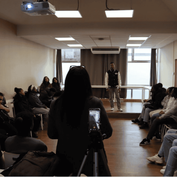 Un trépied avec un téléphone est au premier plan et filme une intervenante de dos et en arrière plan un étudiant sur une estrade. D'autres étudiants sont assis de chaque côté